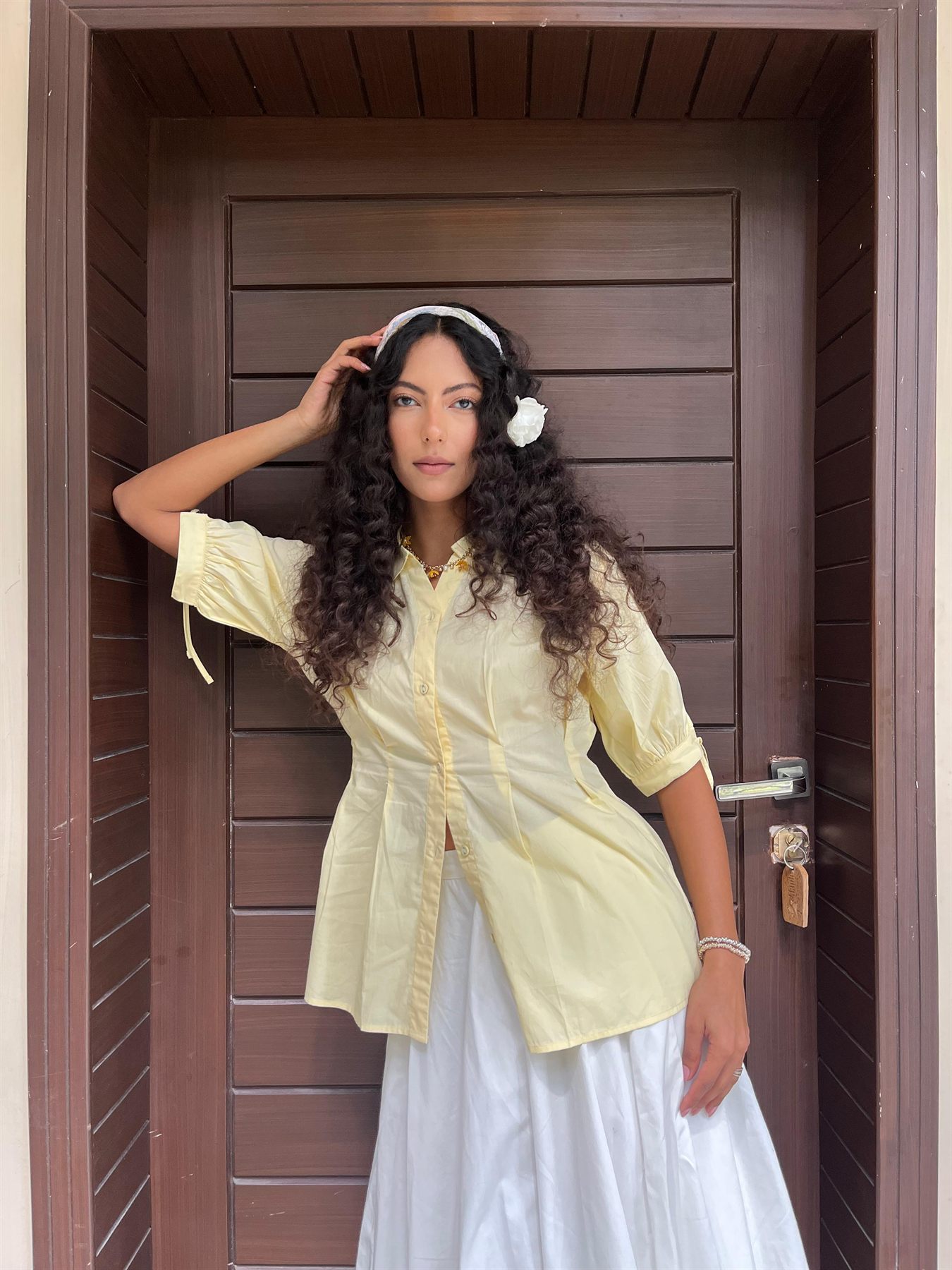 Nimra Jacob in a pale yellow shirt and white skirt against a wooden door